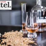 A glass of brown spirit on a table next to some grain