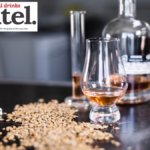 A glass of brown spirit on a table next to some grain