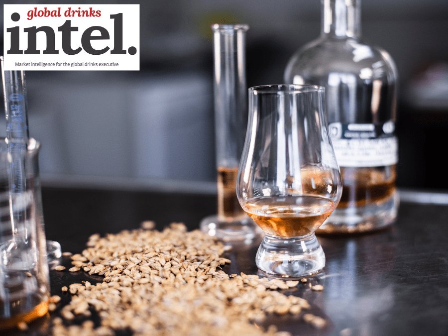 A glass of brown spirit on a table next to some grain