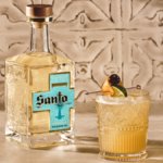 Bottle of Santo Tequila next to a cocktail against stone wall
