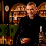 Liev Schreiber at a bar with a bottle of Slainte Irish whiskey