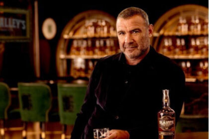 Liev Schreiber at a bar with a bottle of Slainte Irish whiskey