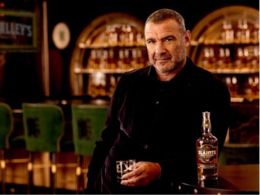 Liev Schreiber at a bar with a bottle of Slainte Irish whiskey