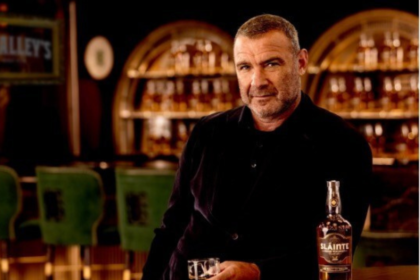 Liev Schreiber at a bar with a bottle of Slainte Irish whiskey