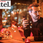 A man at a table in a bar looking at a cellphone with a laptop open