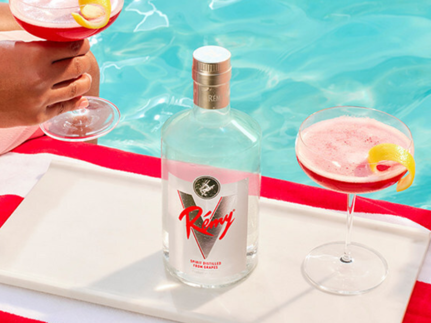 White spirit Remy V on a sun bed with stripey towel and two hands holding red cocktails either side